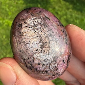 May include: A polished, oval-shaped gemstone with a mix of pink, silver, and black colors. The stone has a textured surface and is held in a person's hand. The background is blurred green grass.