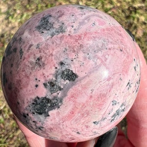 May include: A pink and black polished sphere of rhodochrosite, a type of gemstone. The sphere has a smooth, polished surface and a variety of pink and black patterns.