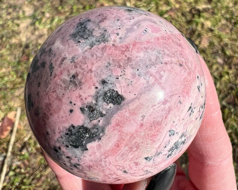May include: A pink and black polished sphere of rhodochrosite, a type of gemstone. The sphere has a smooth, polished surface and a variety of pink and black patterns.