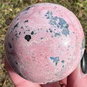 May include: A pink and black sphere of polished rhodochrosite, a type of gemstone. The sphere has a smooth, polished surface with black inclusions throughout.