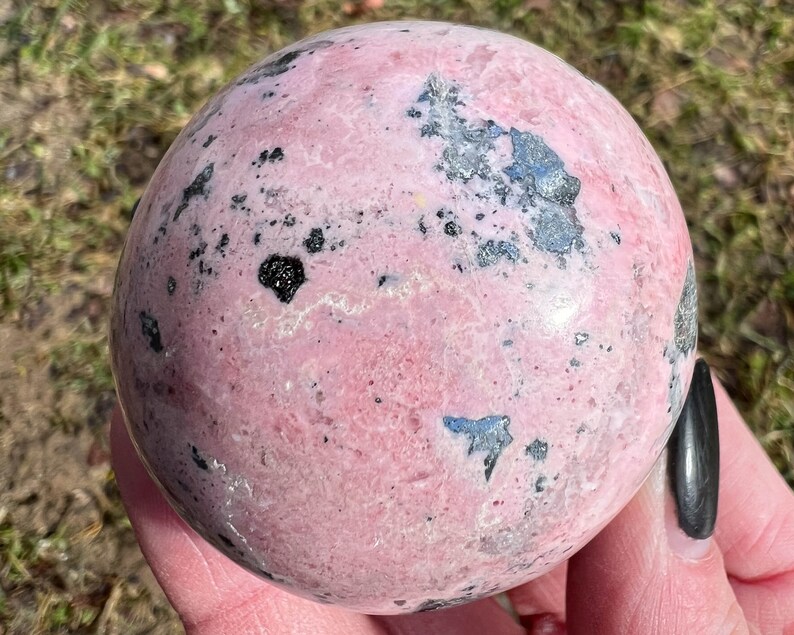 May include: A pink and black sphere of polished rhodochrosite, a type of gemstone. The sphere has a smooth, polished surface with black inclusions throughout.