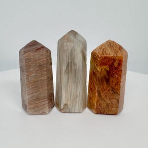 May include: Three polished crystal obelisks. The obelisks are in shades of brown, tan, and orange. Each has a flat top and a rectangular base. The crystals have visible natural patterns and textures. The obelisks are standing upright on a white surface.