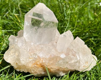May include: A large clear quartz crystal cluster with a prominent point, surrounded by smaller crystals. The cluster is resting on a bed of green grass.