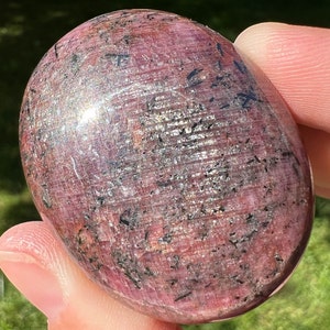 May include: A smooth, oval-shaped pink ruby gemstone with black inclusions. The stone is polished and has a shiny, reflective surface.
