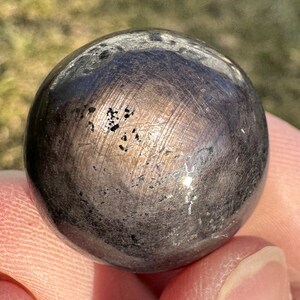 Puede incluir: Una esfera lisa de color gris oscuro con un brillo metálico. La esfera tiene algunas manchas pequeñas y oscuras en su superficie.
