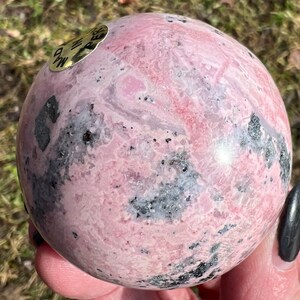May include: A pink and black rhodochrosite sphere with a gold label that reads "Rhodochrosite".