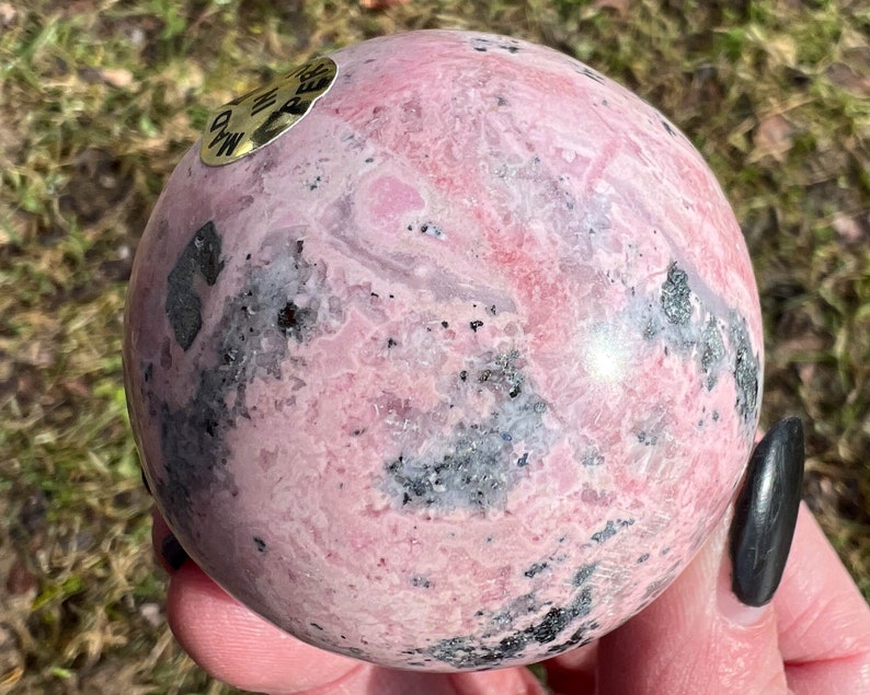 May include: A pink and black rhodochrosite sphere with a gold label that reads "Rhodochrosite".