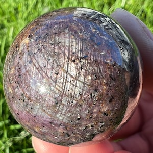 Könnte beinhalten: Eine polierte, dunkelviolette Kugel mit einem einzigartigen, wirbelnden Muster aus silbernen und goldenen Einschlüssen. Die Kugel ist aus natürlichem Edelstein gefertigt und hat eine glatte, reflektierende Oberfläche.