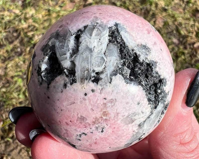 May include: A pink and white sphere with black inclusions. The sphere is polished and has a smooth surface. It is a natural crystal and has a unique pattern.