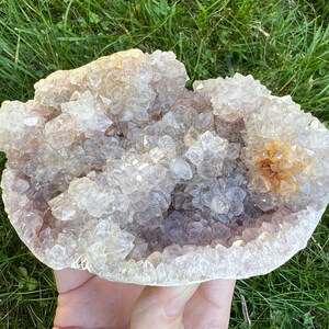K&ouml;nnte beinhalten: Ein gro&szlig;er, klarer Quarzkristall-Cluster mit einer nat&uuml;rlichen, drusigen Oberfl&auml;che. Der Kristall hat eine hellviolette Farbe und einen kleinen, orangebraunen Kristall-Cluster in der Mitte.
