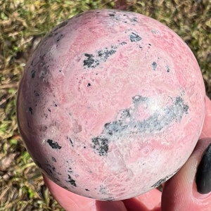 May include: A pink and black polished sphere of rhodochrosite crystal. The sphere has a smooth, polished surface with a swirling pattern of pink and black.