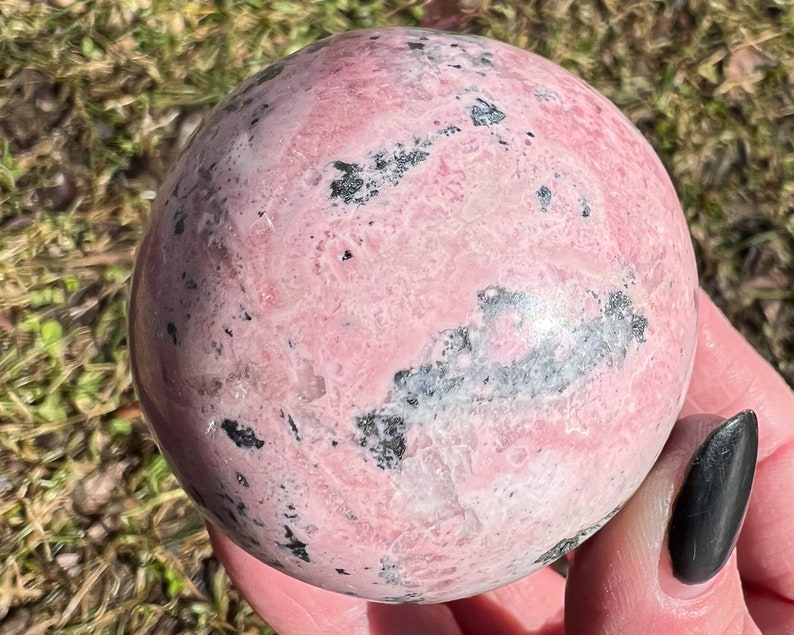 May include: A pink and black polished sphere of rhodochrosite crystal. The sphere has a smooth, polished surface with a swirling pattern of pink and black.