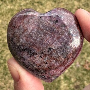Puede incluir: Un cristal de rubí pulido en forma de corazón, de color rojo intenso y morado. El cristal tiene una superficie lisa y pulida y una forma natural y orgánica.