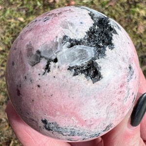 May include: A pink and white sphere with black inclusions. The sphere is made of polished stone and has a smooth, glossy finish.