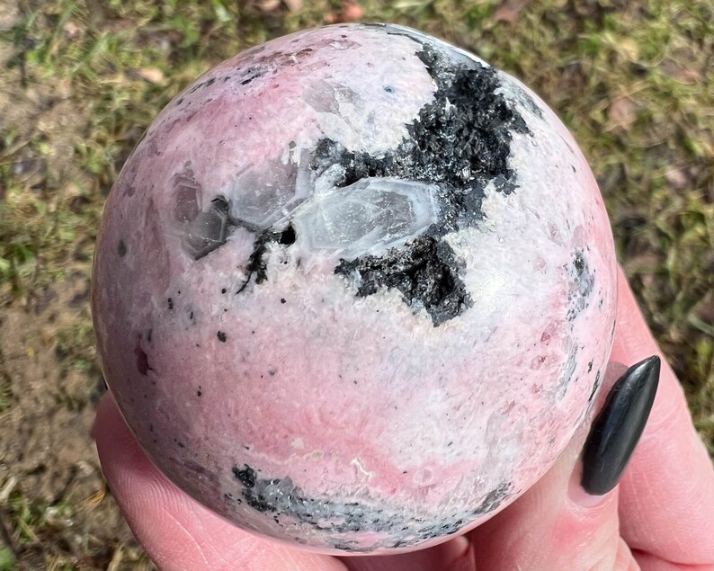 May include: A pink and white sphere with black inclusions. The sphere is made of polished stone and has a smooth, glossy finish.