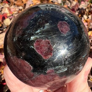 May include: A black sphere with red and silver inclusions. The sphere is polished and has a reflective surface.