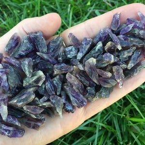 May include: A hand holding a pile of amethyst crystal points. The crystals are a mix of purple and green hues and have a natural, raw look.