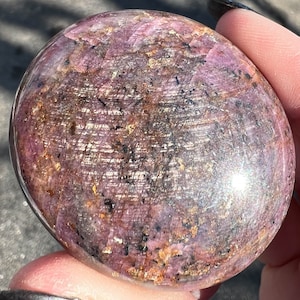 May include: A polished pink and brown gemstone with a smooth, rounded surface. The stone has a sparkly, iridescent finish.