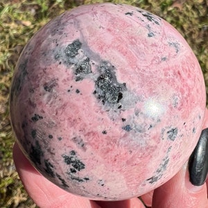 May include: A pink and black sphere of polished rhodochrosite, a type of gemstone. The sphere has a smooth, polished surface and a swirling pattern of pink and black.