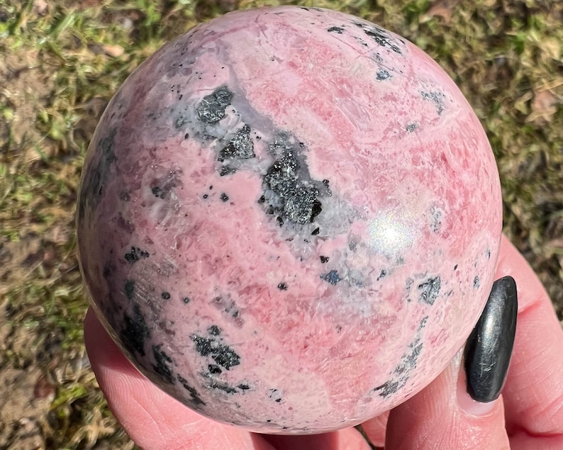 May include: A pink and black sphere of polished rhodochrosite, a type of gemstone. The sphere has a smooth, polished surface and a swirling pattern of pink and black.