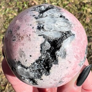 May include: A pink and black sphere of polished Rhodonite with white Quartz inclusions. The sphere is smooth and polished, with a natural, organic look.
