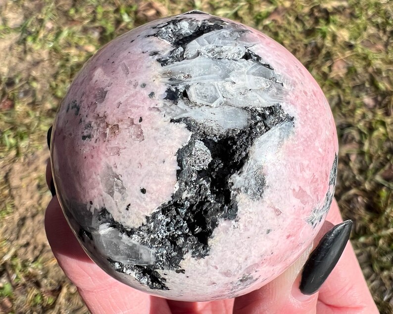 May include: A pink and black sphere of polished Rhodonite with white Quartz inclusions. The sphere is smooth and polished, with a natural, organic look.