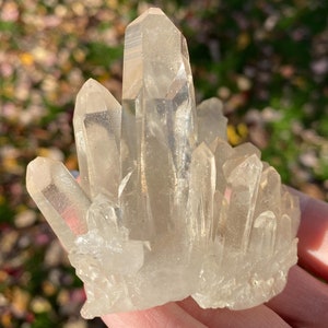 Puede incluir: Un c&uacute;mulo de cristales de cuarzo transparentes de diferentes tama&ntilde;os y formas. Los cristales est&aacute;n dispuestos en un c&uacute;mulo, con algunos de los cristales apuntando hacia arriba y otros hacia abajo. Los cristales son transl&uacute;cidos y tienen un ligero brillo.