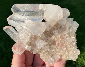 May include: A cluster of clear quartz crystals with a base of smaller, milky quartz crystals. The larger crystals are clear and have a glassy finish. The smaller crystals are opaque and have a milky white color.