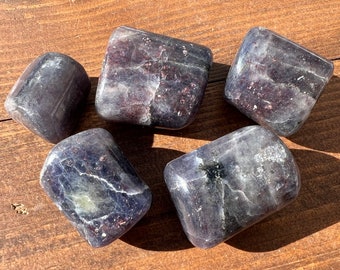 May include: Five tumbled stones of purple and gray amethyst, with a smooth, polished finish. The stones are arranged on a wooden surface.