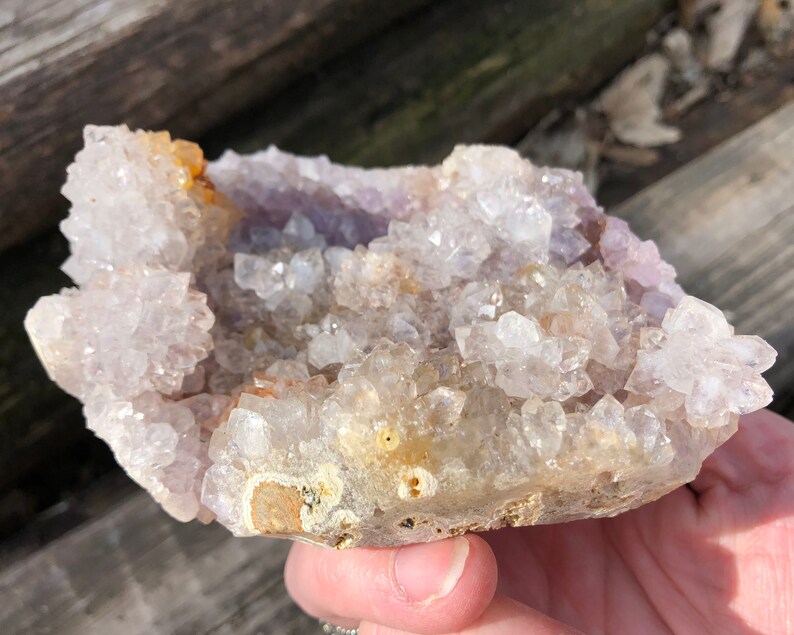 K&ouml;nnte beinhalten: Ein Cluster aus klaren Quarzkristallen mit einem violetten Amethyst-Zentrum. Die Kristalle sind in einer nat&uuml;rlichen Formation angeordnet, wobei einige der Spitzen nach oben zeigen. Der Cluster ist etwa 12,5 cm breit und 7,5 cm hoch.