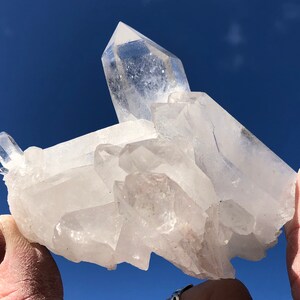 May include: A large, clear quartz crystal cluster with multiple points. The crystal is translucent and has a natural, rough texture. The cluster is held in a hand against a blue sky background.