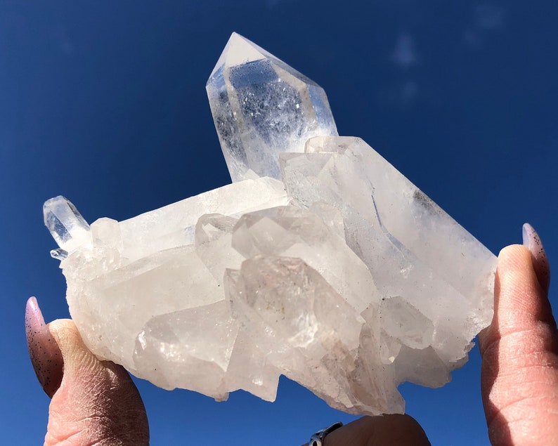 May include: A large, clear quartz crystal cluster with multiple points. The crystal is translucent and has a natural, rough texture. The cluster is held in a hand against a blue sky background.