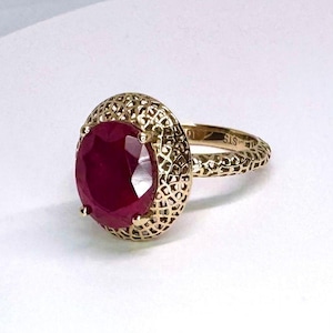 May include: A gold ring featuring a large, oval-cut ruby gemstone. The ruby is a deep red color and is set in a textured, filigree-style gold setting. The band of the ring also has a textured design.