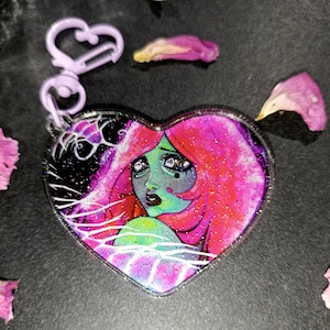 Può includere: Portachiavi a forma di cuore con un'illustrazione colorata di una donna con capelli rosa e pelle verde. L'opera d'arte ha una finitura glitterata ed è attaccata a un fermaglio color lavanda. Lo sfondo è scuro con linee bianche.