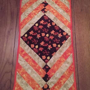 May include: A quilted table runner with a diamond pattern in the center. The diamond is made of a fabric with a brown background and a pattern of orange and yellow leaves. The runner is surrounded by a border of orange and white stripes.