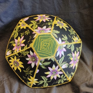 Könnte beinhalten: Eine schwarze Stoffschale mit geometrischem Muster. Die Schale ist mit einem floralen Muster aus lila und gelben Blumen auf schwarzem Hintergrund verziert. Die Schale ist mit goldenen Verzierungen eingefasst.