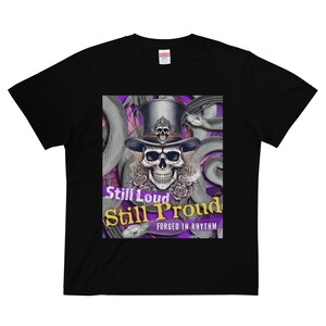 Può includere: T-shirt nera con un teschio con un cappello a cilindro, circondato da serpenti e rose. Il testo "Still Loud Still Proud" e "Forged in Rhythm" è visualizzato in un design viola e oro.