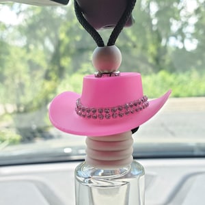 May include: A pink cowboy hat car air freshener with a clear glass bottle. The hat is adorned with a row of sparkling rhinestones. The air freshener hangs from a black cord and a wooden bead.
