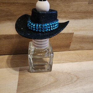 May include: A black glitter cowboy hat with a turquoise rhinestone band sits atop a clear glass bottle. The hat is attached to a black cord with a white ball. The bottle is filled with a clear liquid.