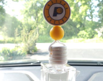 May include: Car air freshener with an Indiana Pacers logo. The air freshener has a clear, square-shaped bottle, a white top, a yellow sphere, and a circular logo with the team's logo and the words "INDIANA PACERS".