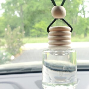 K&ouml;nnte beinhalten: Klarer Auto-Lufterfrischer-Flasche aus Glas mit einem Holzdeckel und einer Holzperle, die an einer schwarzen Schnur h&auml;ngt. Die Flasche ist mit einer klaren Fl&uuml;ssigkeit gef&uuml;llt. Der Hintergrund ist eine verschwommene Ansicht von B&auml;umen und einer Stra&szlig;e.
