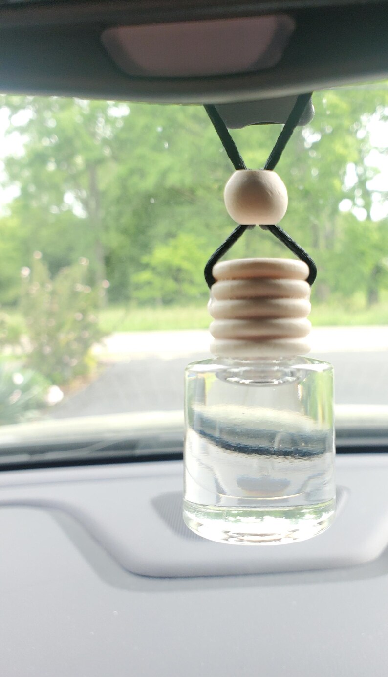 K&ouml;nnte beinhalten: Klarer Auto-Lufterfrischer-Flasche aus Glas mit einem Holzdeckel und einer Holzperle, die an einer schwarzen Schnur h&auml;ngt. Die Flasche ist mit einer klaren Fl&uuml;ssigkeit gef&uuml;llt. Der Hintergrund ist eine verschwommene Ansicht von B&auml;umen und einer Stra&szlig;e.
