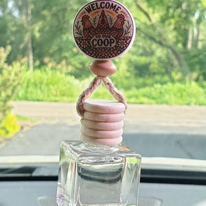 May include: A clear, square glass bottle with a wooden top and a decorative charm. The charm features the text "WELCOME TO OUR COOP" around a design of chickens. The item is suspended by a brown rope.