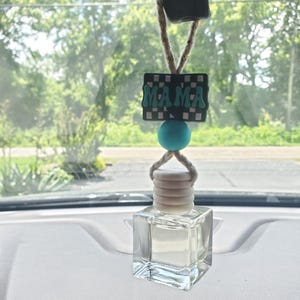 May include: Car air freshener with a clear, square glass bottle and a white cap. The freshener hangs from a string and features a black and white checkered rectangle with the word "MAMA" in teal letters, and a teal bead.