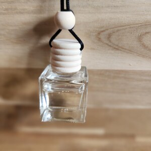 K&ouml;nnte beinhalten: Eine klare, quadratische Glasflasche mit einem Holzverschluss und einer schwarzen Schnur zum Aufh&auml;ngen. Die Flasche ist mit einer klaren Fl&uuml;ssigkeit gef&uuml;llt. &Uuml;ber dem Verschluss befindet sich eine Holzperle. Der Hintergrund ist eine hellbraune Holzoberfl&auml;che.