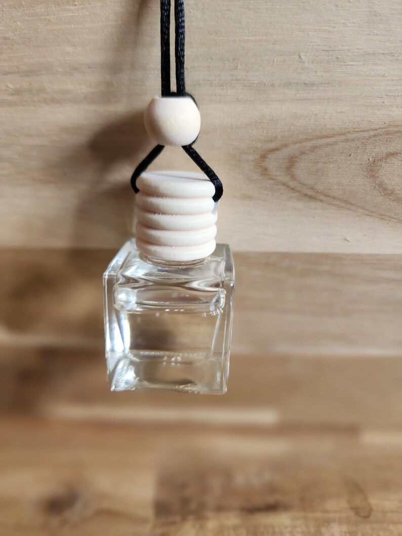 K&ouml;nnte beinhalten: Eine klare, quadratische Glasflasche mit einem Holzverschluss und einer schwarzen Schnur zum Aufh&auml;ngen. Die Flasche ist mit einer klaren Fl&uuml;ssigkeit gef&uuml;llt. &Uuml;ber dem Verschluss befindet sich eine Holzperle. Der Hintergrund ist eine hellbraune Holzoberfl&auml;che.