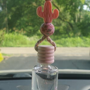 May include: A clear glass car air freshener with a pink cactus charm and a pink and gold beaded accent. The air freshener is hanging from a brown string.