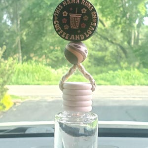 May include: A car air freshener with a clear glass bottle and a wooden top. Above the bottle is a decorative piece with the text "THIS MAMA RUNS ON COFFEE AND LOVE" and a coffee cup graphic. The air freshener hangs from a string.