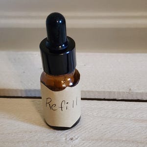 May include: A small amber glass bottle with a black dropper top. A label with the word "Refill" is affixed to the bottle. The bottle is sitting on a white wooden surface.