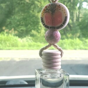 May include: A car air freshener featuring a clear, square glass bottle and a light pink wooden top. A round charm with the text "JUST A GIRL WHO LOVES BOOKS" and a floral book design hangs above the bottle. The charm is connected by a brown string.